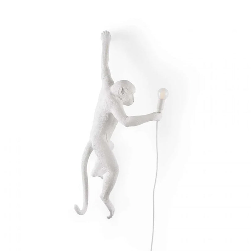 A white Clérachic monkey wall light on a white backdrop, its raised arm and long tail leaving a faint shadow on the wall.
