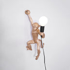 A gold Clérachic monkey wall light facing right, its raised arm gripping the wall and a globe bulb held at its side.