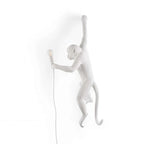 A white Clérachic monkey light, facing left, hangs by one arm on a white backdrop with a small bulb held at its side.