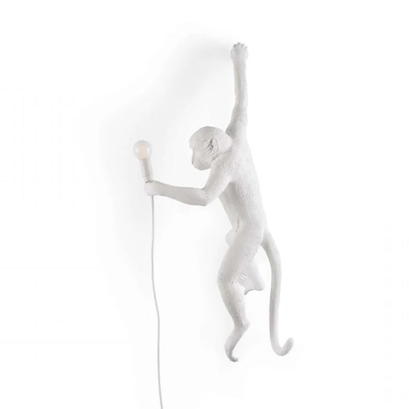 A white Clérachic monkey light, facing left, hangs by one arm on a white backdrop with a small bulb held at its side.
