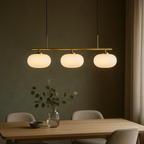 Clérachic three-head glass pendant light in a gold-toned finish, lit above a table set with a glass vase of eucalyptus branches against a deep green wall.