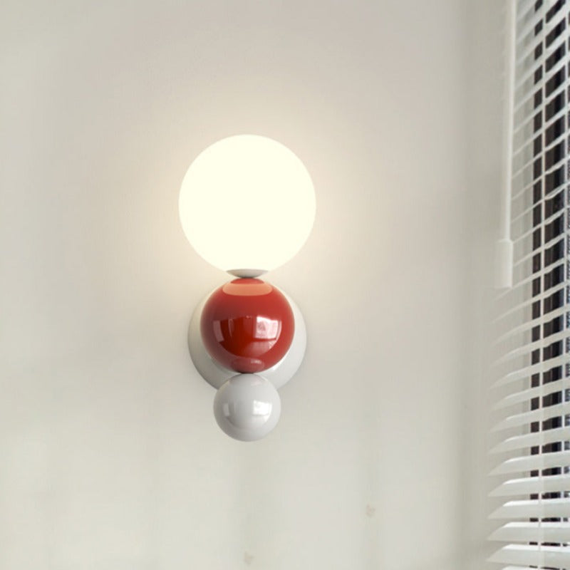 The Clérachic three-sphere wall light in red, a glossy red sphere between the lit opal globe and a white base sphere, beside window blinds.