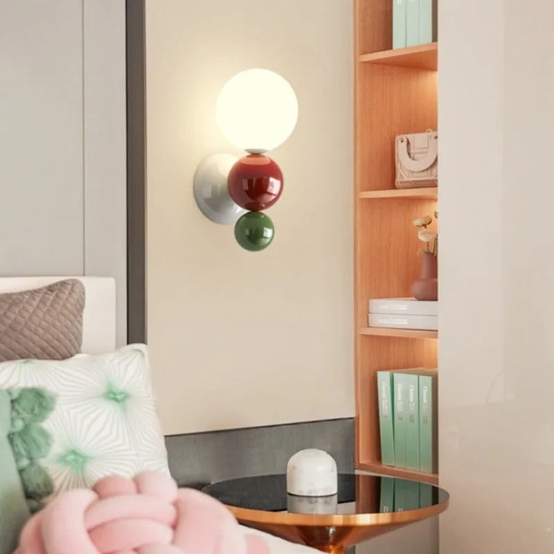 The Clérachic three-sphere wall light in green, lit beside a wooden bookshelf with plants and books in a reading nook.
