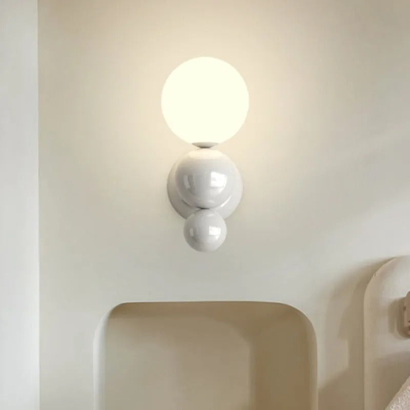 The Clérachic three-sphere wall light in white, glowing above a curved headboard nook in a bedroom.