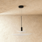 Clérachic pendant with two stacked white frosted discs and a black weight on thin wires, on a limewash backdrop.
