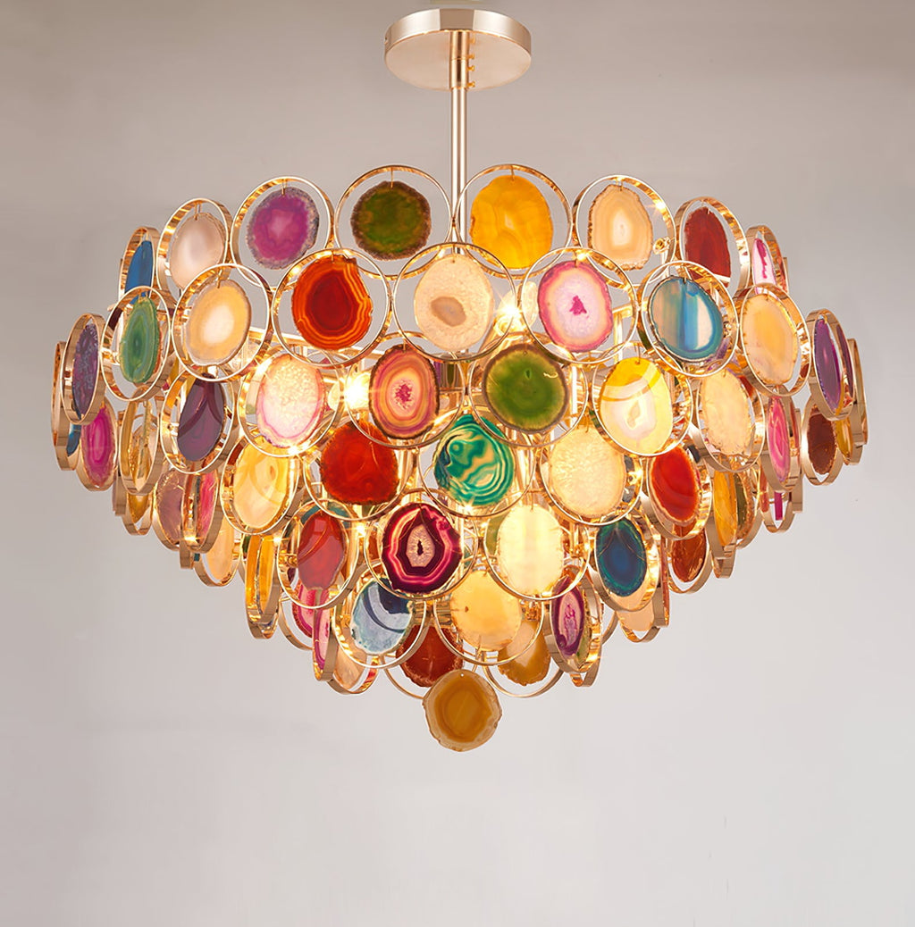 A Clérachic agate chandelier on a pale grey wall, its stacked tiers of coloured stone slices glowing with warm light.