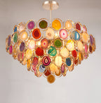 A Clérachic agate chandelier on a pale grey wall, its stacked tiers of coloured stone slices glowing with warm light.