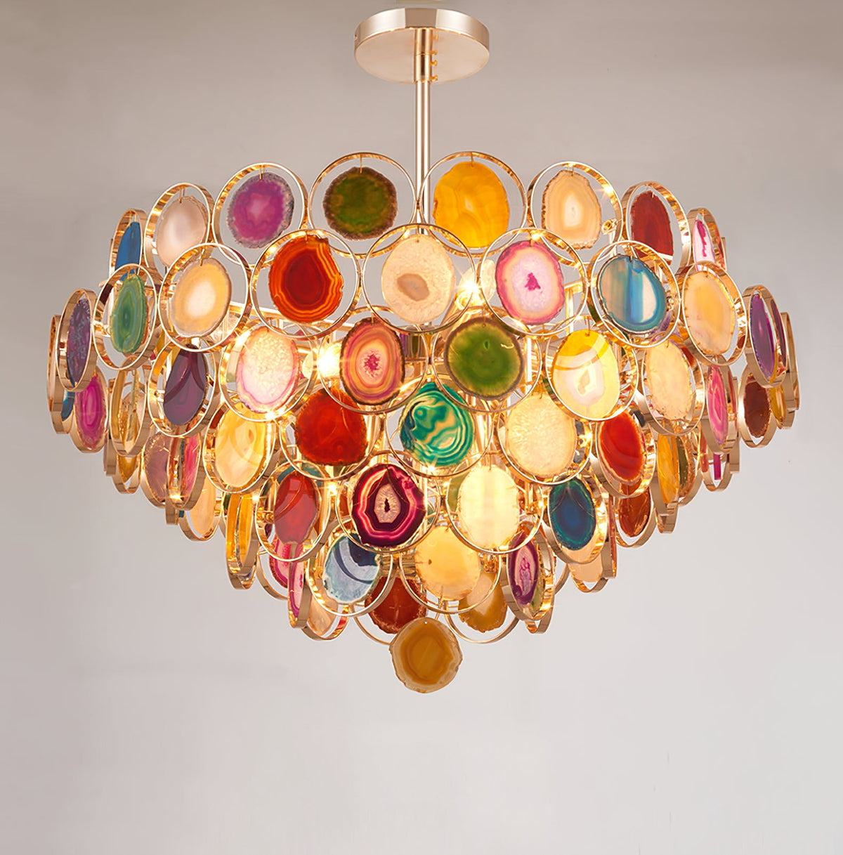 A Clérachic agate chandelier on a pale grey wall, its stacked tiers of coloured stone slices glowing with warm light.