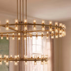 Close view of a Clérachic lacquered brass chandelier lit warm, its glass cylinders glowing on two rings in a dining room.