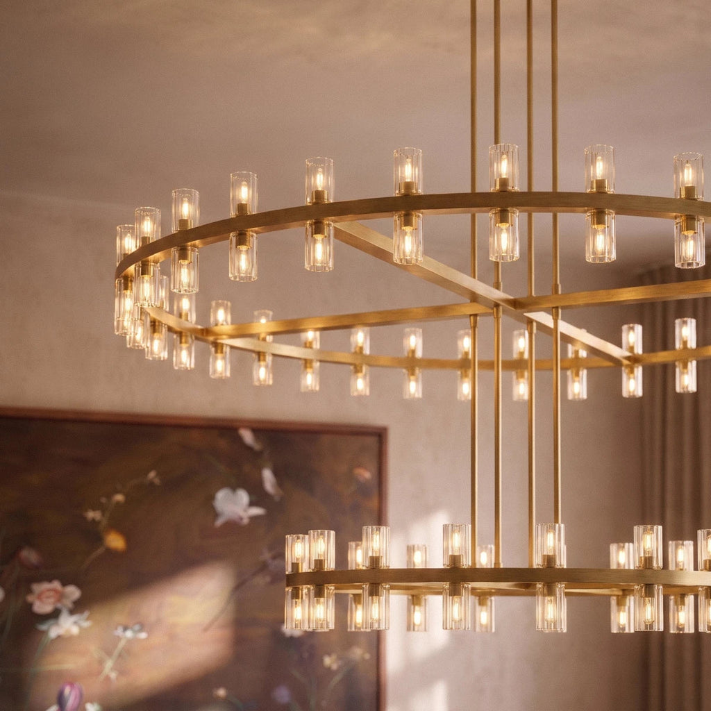 Close view of a Clérachic lacquered brass chandelier with lit glass cylinders, seen against a floral painting and linen curtains.