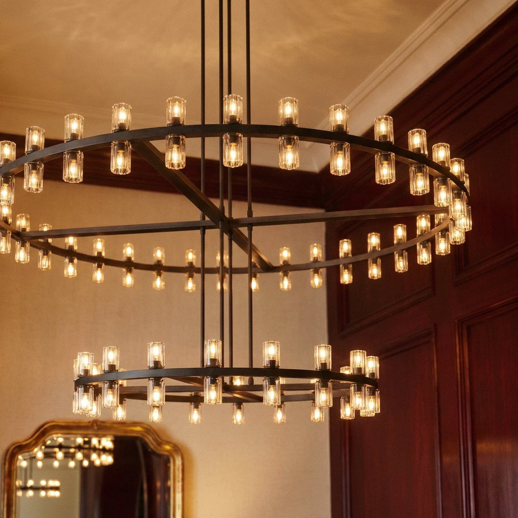 Close view of a Clérachic matte black chandelier lit warm, with glass cylinders on both rings beside dark wood panelling.