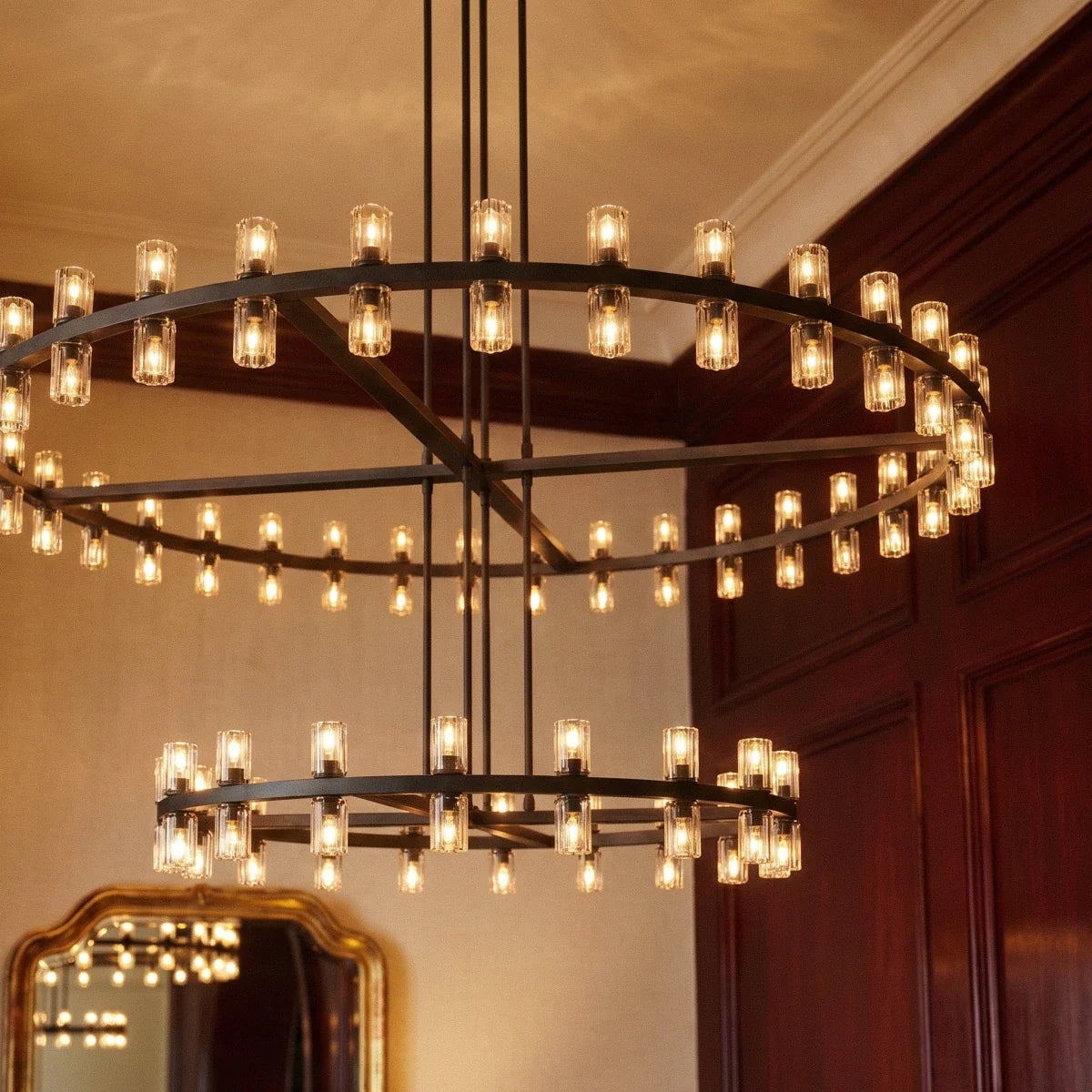 Close view of a Clérachic matte black chandelier lit warm, with glass cylinders on both rings beside dark wood panelling.
