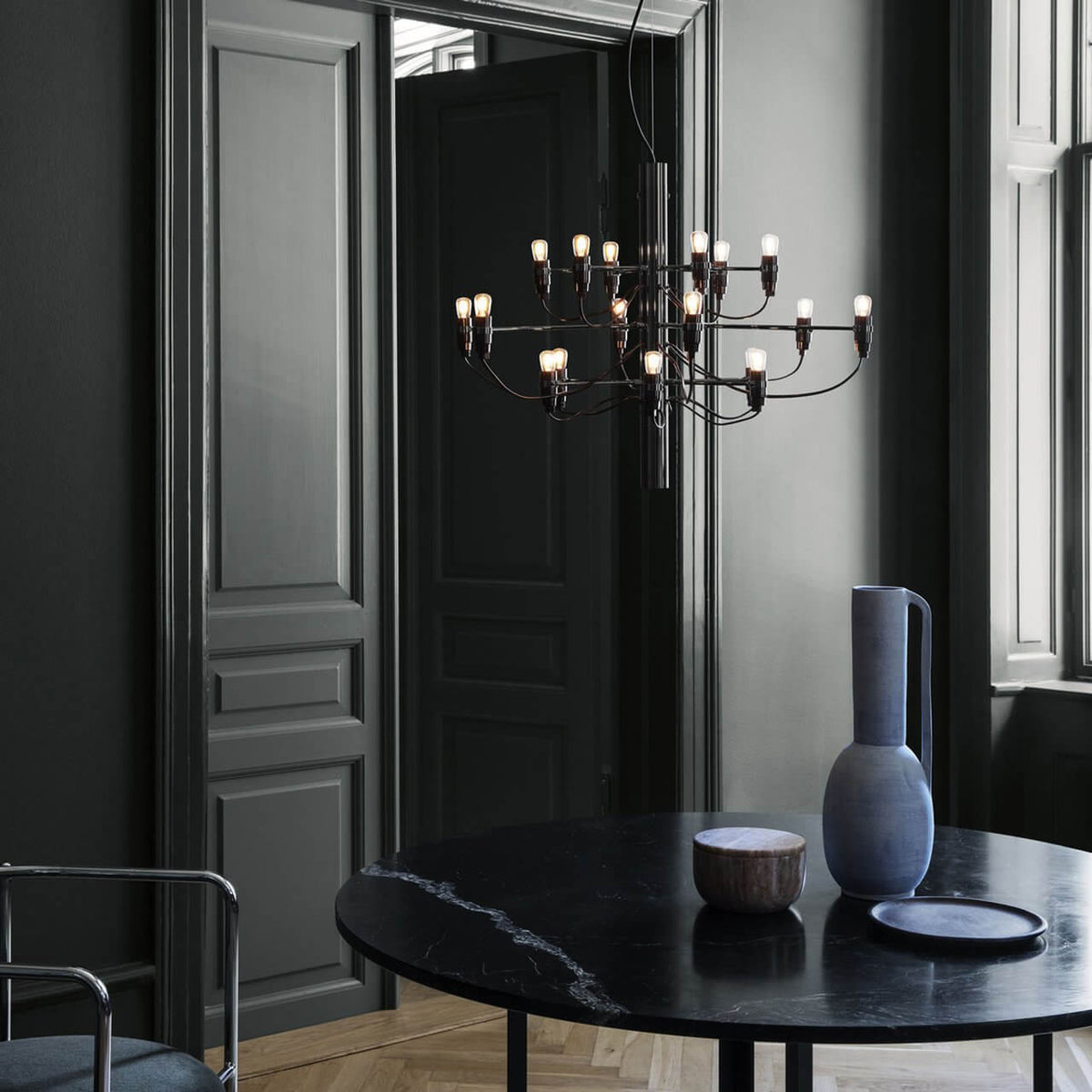 Warm bulbs light this Clérachic chandelier in front of dark panelled doors, above a black marble table and a blue vase.