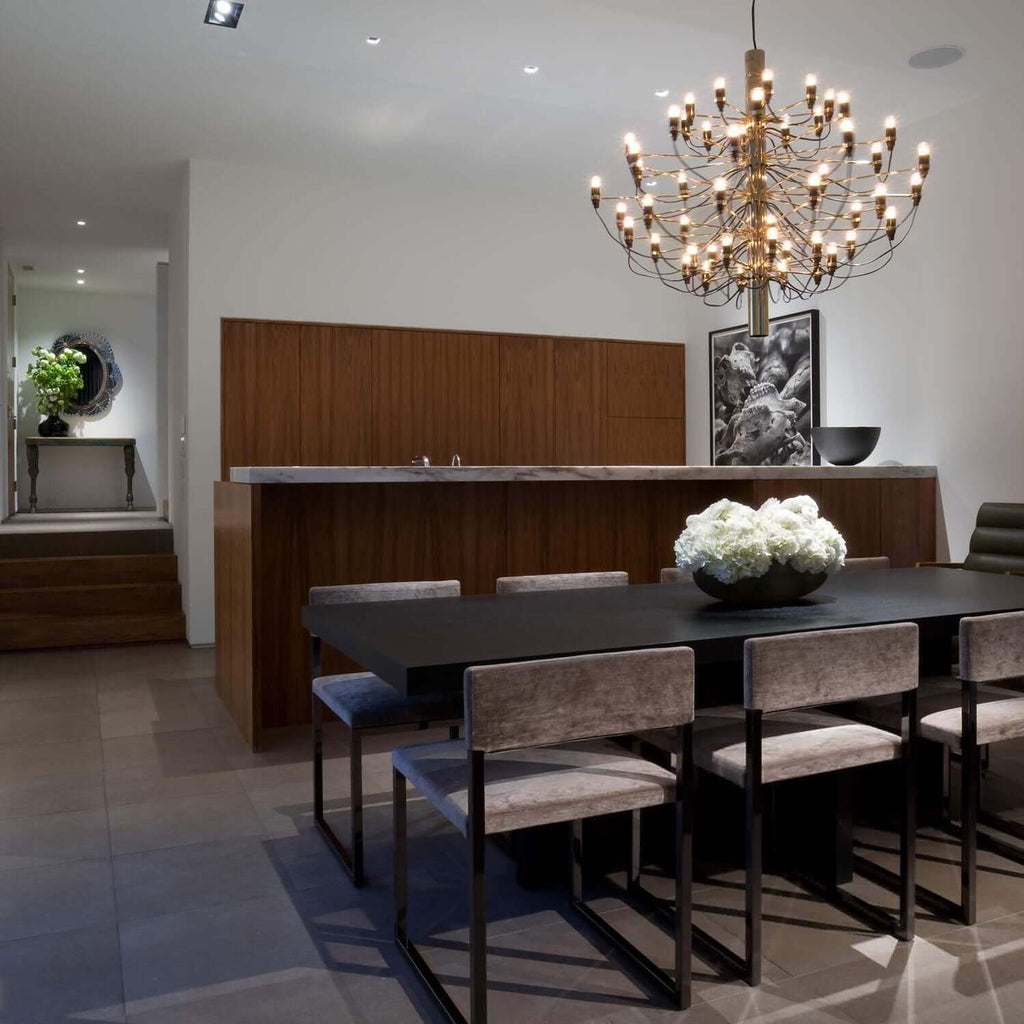 A gold Clérachic chandelier hangs above a dark dining table with grey velvet chairs, in front of a walnut wall.