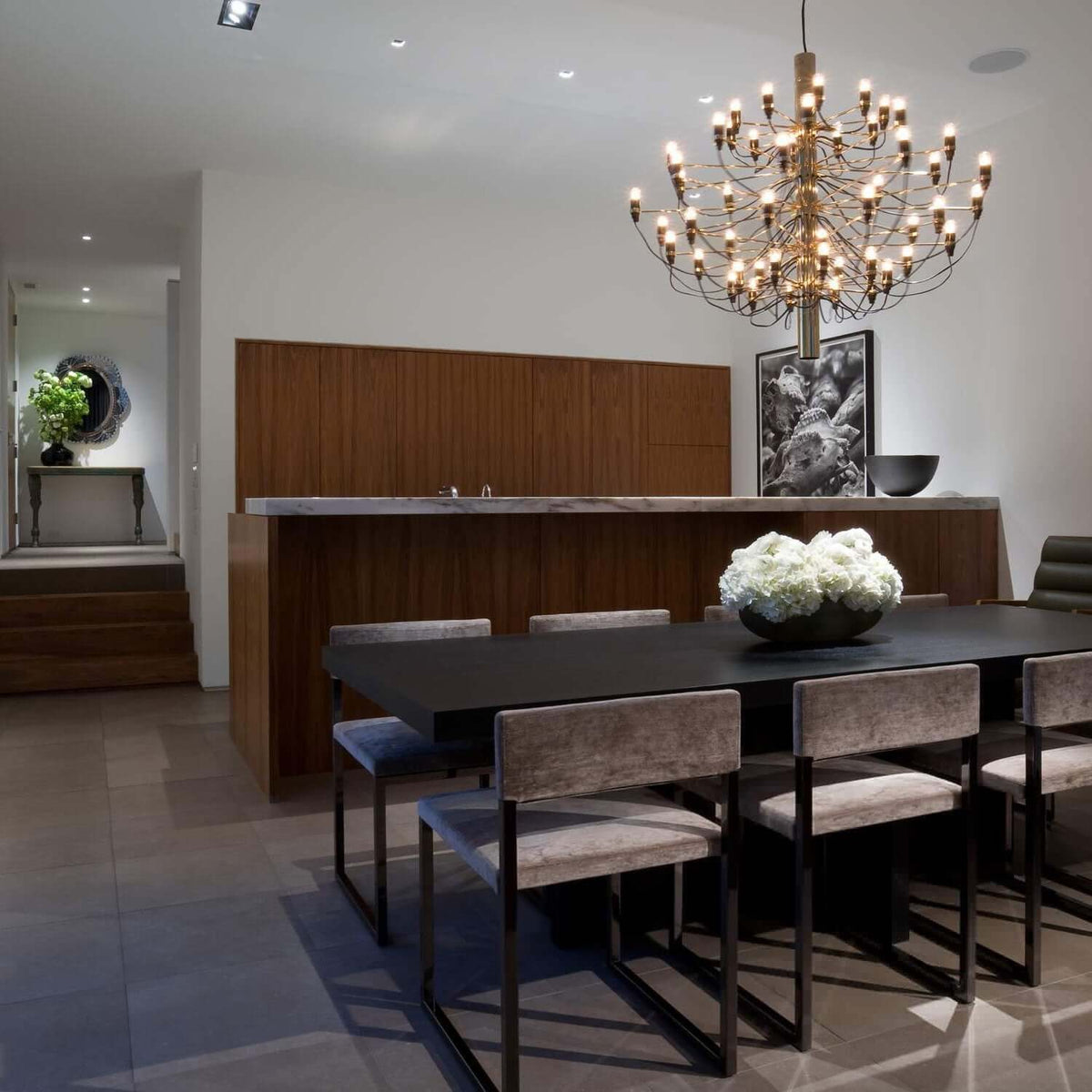 A gold Clérachic chandelier hangs above a dark dining table with grey velvet chairs, in front of a walnut wall.