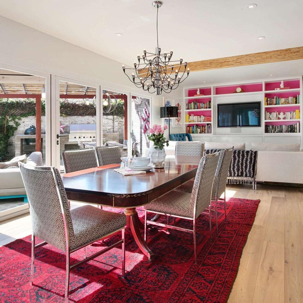 A Clérachic chandelier with a chrome stem hangs over a red rug and a dark dining table, with a pink-backed bookcase behind.