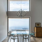 Hung in a double-height room with a glass wall, this Clérachic chandelier lights a table of clear chairs and a sea view.