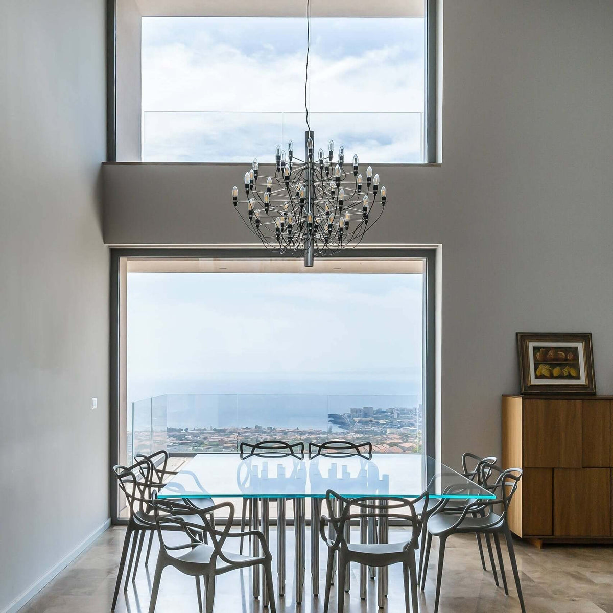 Hung in a double-height room with a glass wall, this Clérachic chandelier lights a table of clear chairs and a sea view.