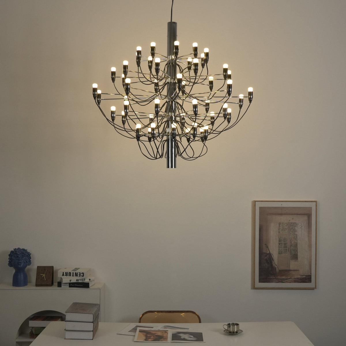 This lit Clérachic chandelier fills a warm-toned wall with soft light, above a desk with books and a framed print.