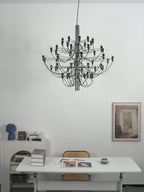 A Clérachic chandelier with a chrome stem hangs against a white wall above a white desk, a blue vase and a framed print.