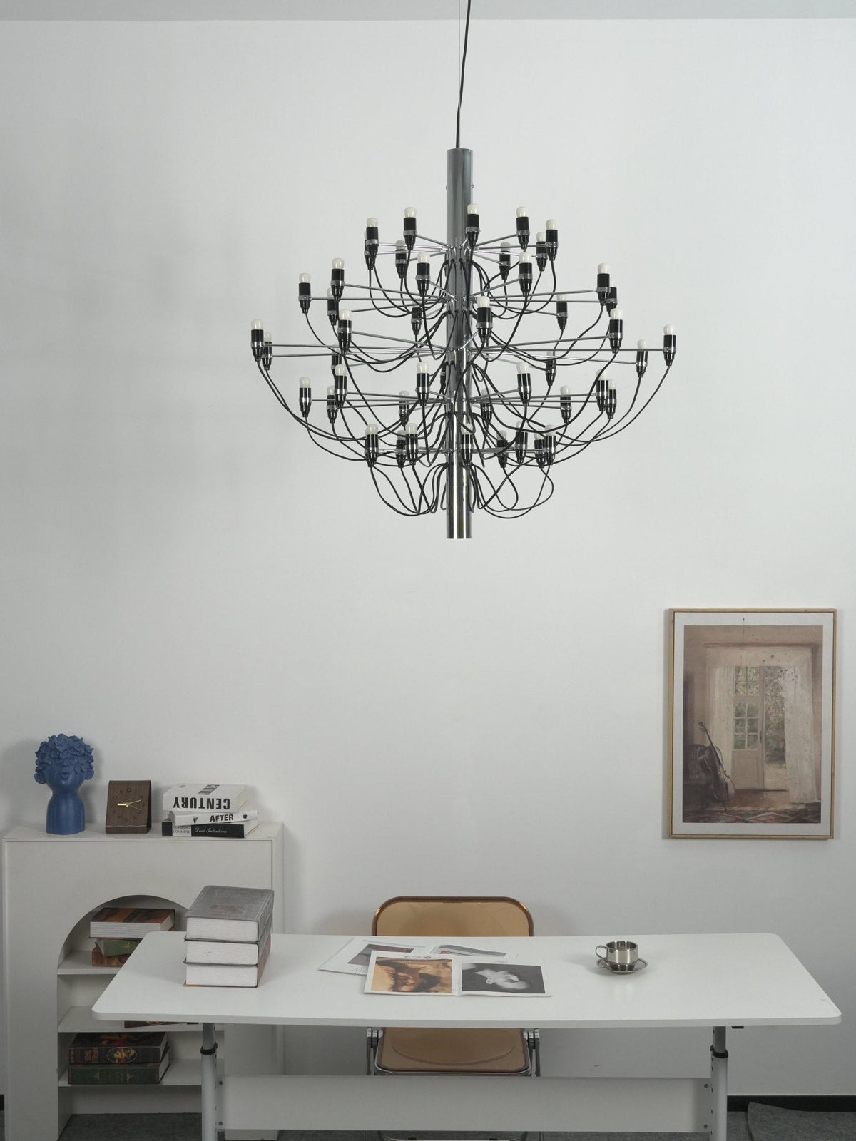 A Clérachic chandelier with a chrome stem hangs against a white wall above a white desk, a blue vase and a framed print.