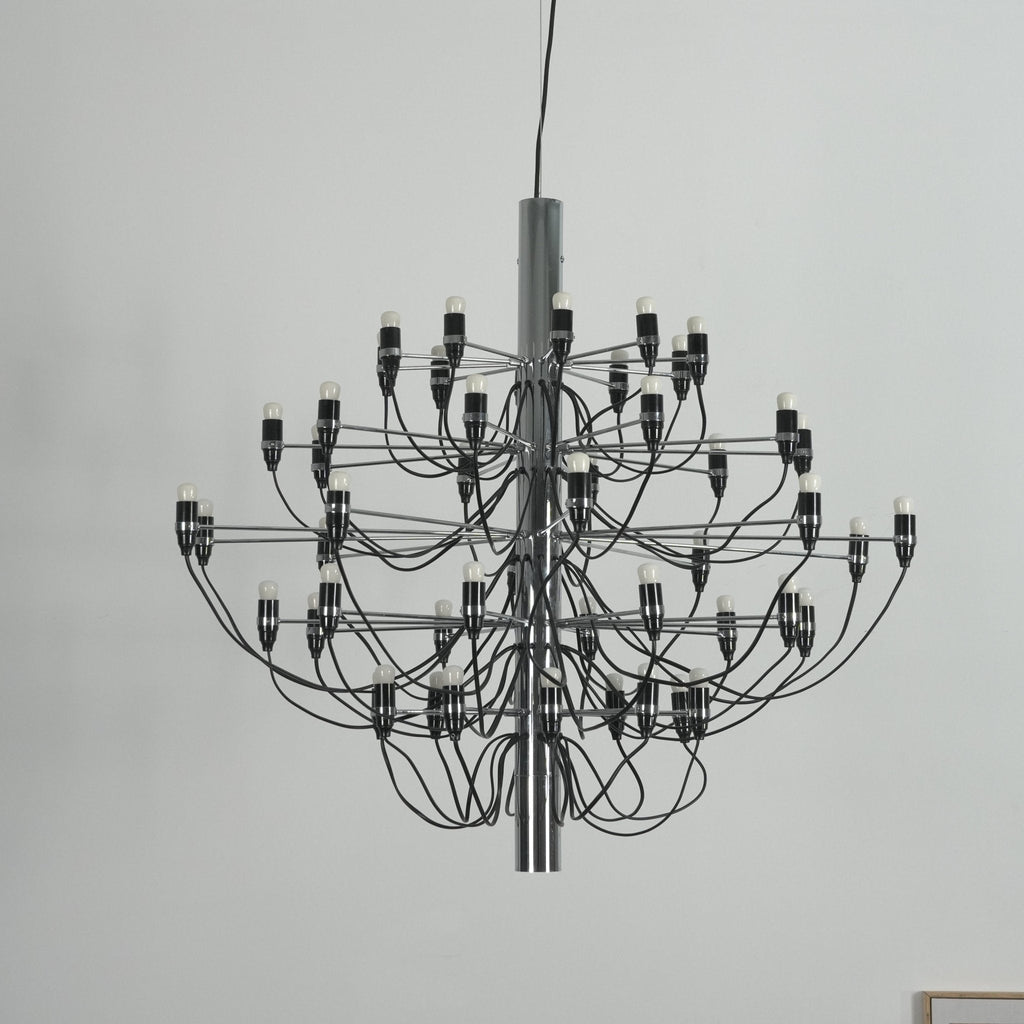 Close view of a Clérachic chandelier with a chrome stem, black cable arms and exposed bulbs against a pale wall.