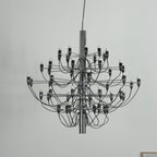 Close view of a Clérachic chandelier with a chrome stem, black cable arms and exposed bulbs against a pale wall.