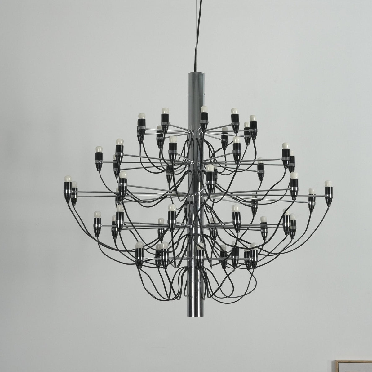 Close view of a Clérachic chandelier with a chrome stem, black cable arms and exposed bulbs against a pale wall.