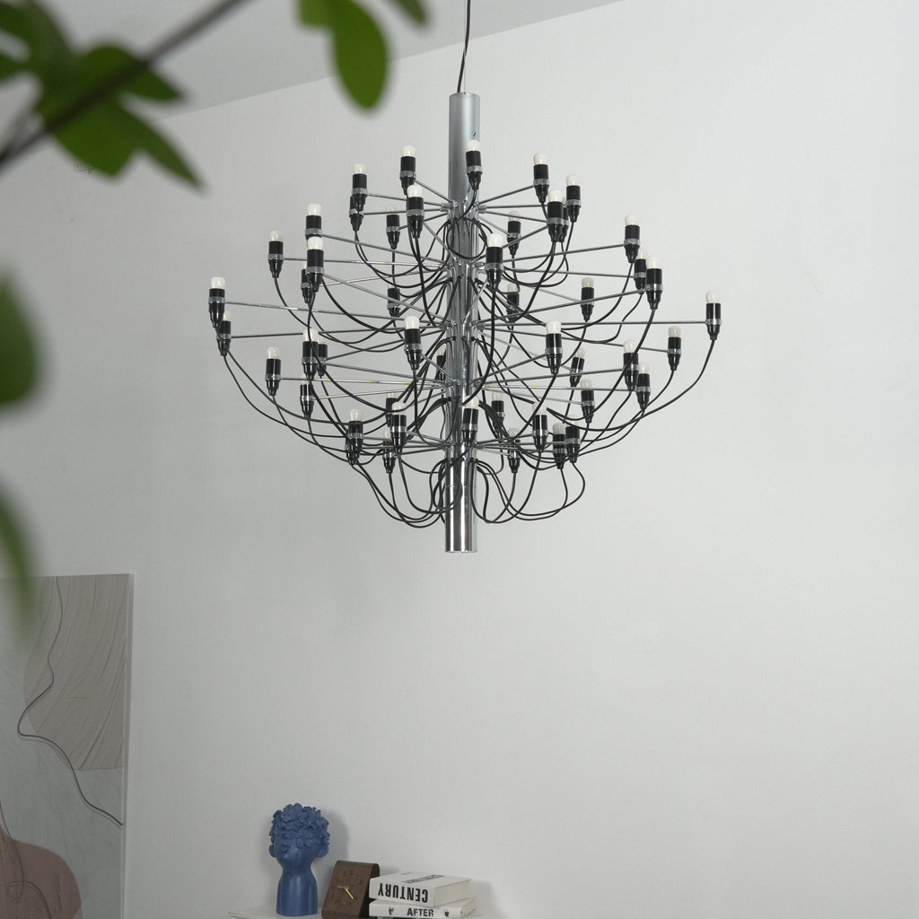 A Clérachic chandelier with a chrome stem hangs beside the leaves of a plant, in front of a white wall and a blue vase.