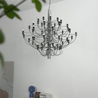 A Clérachic chandelier with a chrome stem hangs beside the leaves of a plant, in front of a white wall and a blue vase.