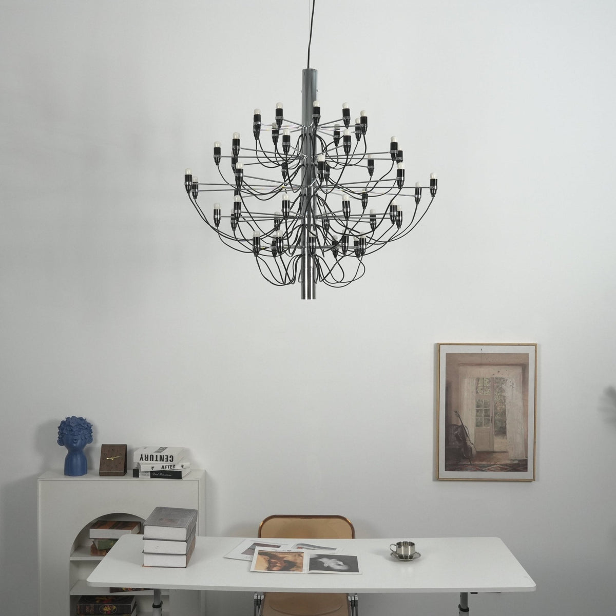 This Clérachic chandelier hangs against a white wall above a desk with books, a blue vase and a framed print.