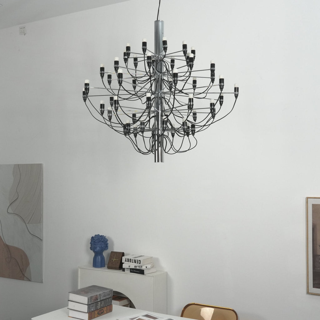 A Clérachic chandelier with a silver-toned stem hangs in a white room, with a leaning art panel and a desk below.