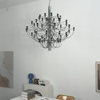 A Clérachic chandelier with a silver-toned stem hangs in a white room, with a leaning art panel and a desk below.
