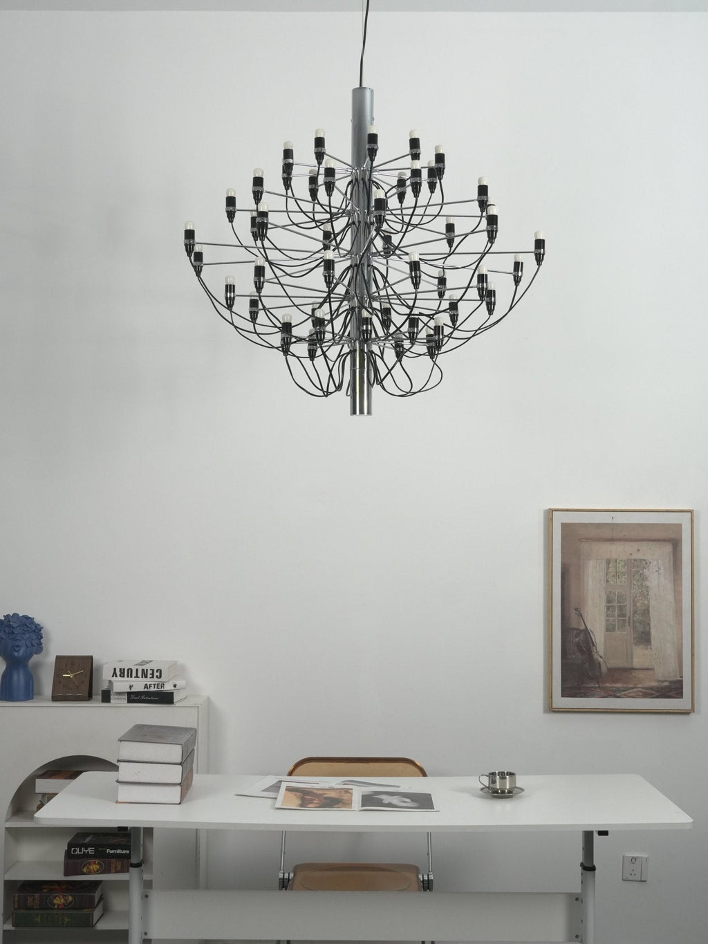 A Clérachic chandelier with slim arms hangs over a white desk, with a woven chair, books and a framed print behind.
