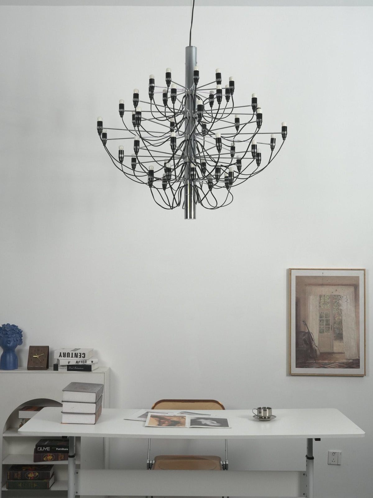A Clérachic chandelier with slim arms hangs over a white desk, with a woven chair, books and a framed print behind.