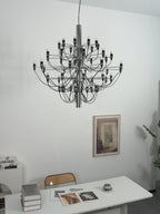 A Clérachic chandelier with a chrome stem hangs close to a white wall, above a desk with magazines and a woven chair.
