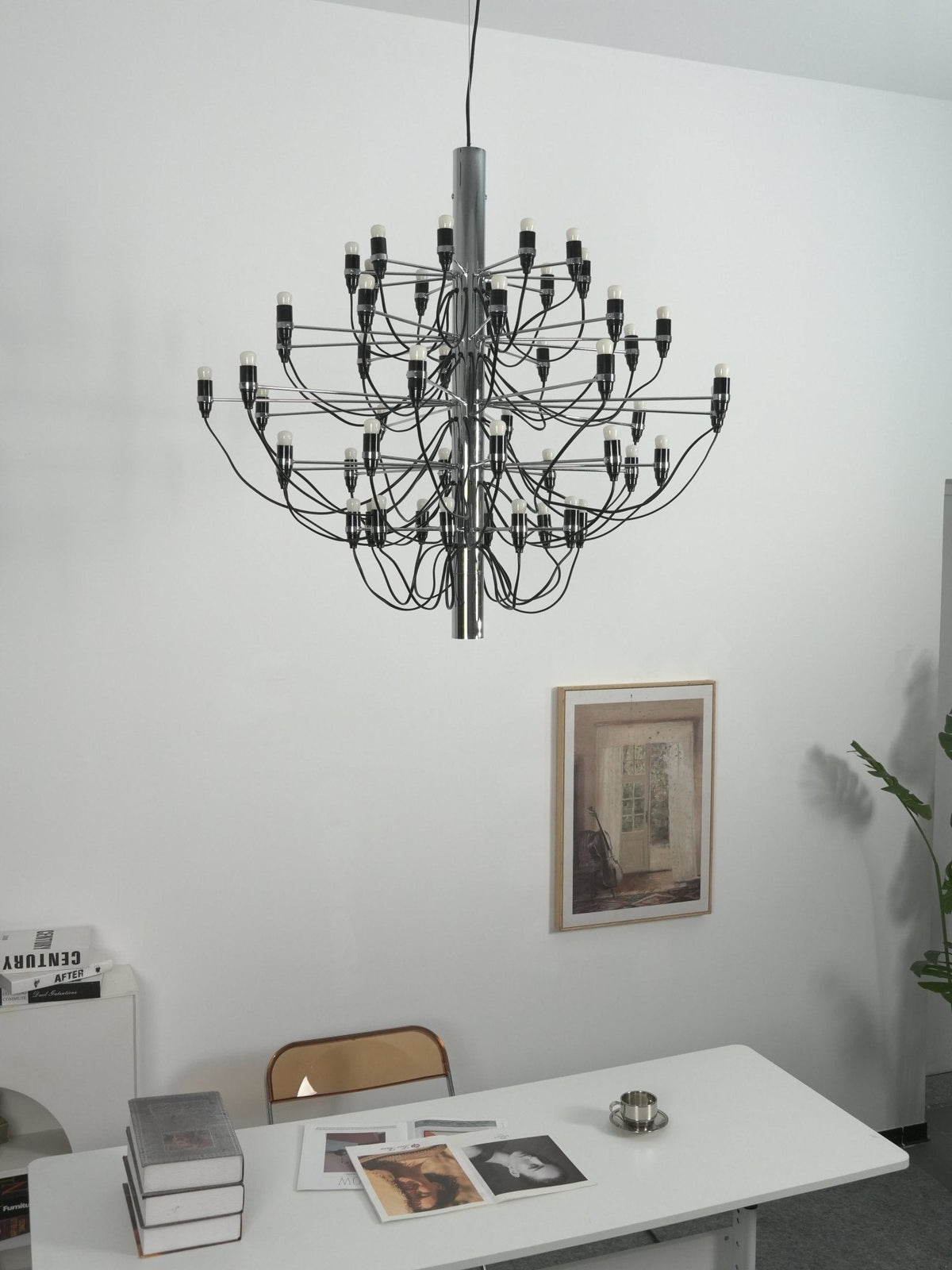 A Clérachic chandelier with a chrome stem hangs close to a white wall, above a desk with magazines and a woven chair.