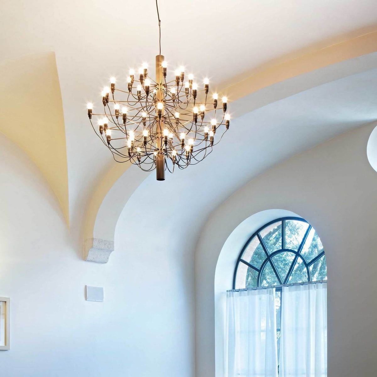 A bronze-toned Clérachic tiered chandelier lit with exposed bulbs, hanging in a vaulted white room beside an arched window.
