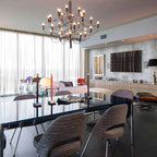 This Clérachic chandelier lights a long glossy dining table in an open-plan room, with sheer curtains and a sideboard behind.