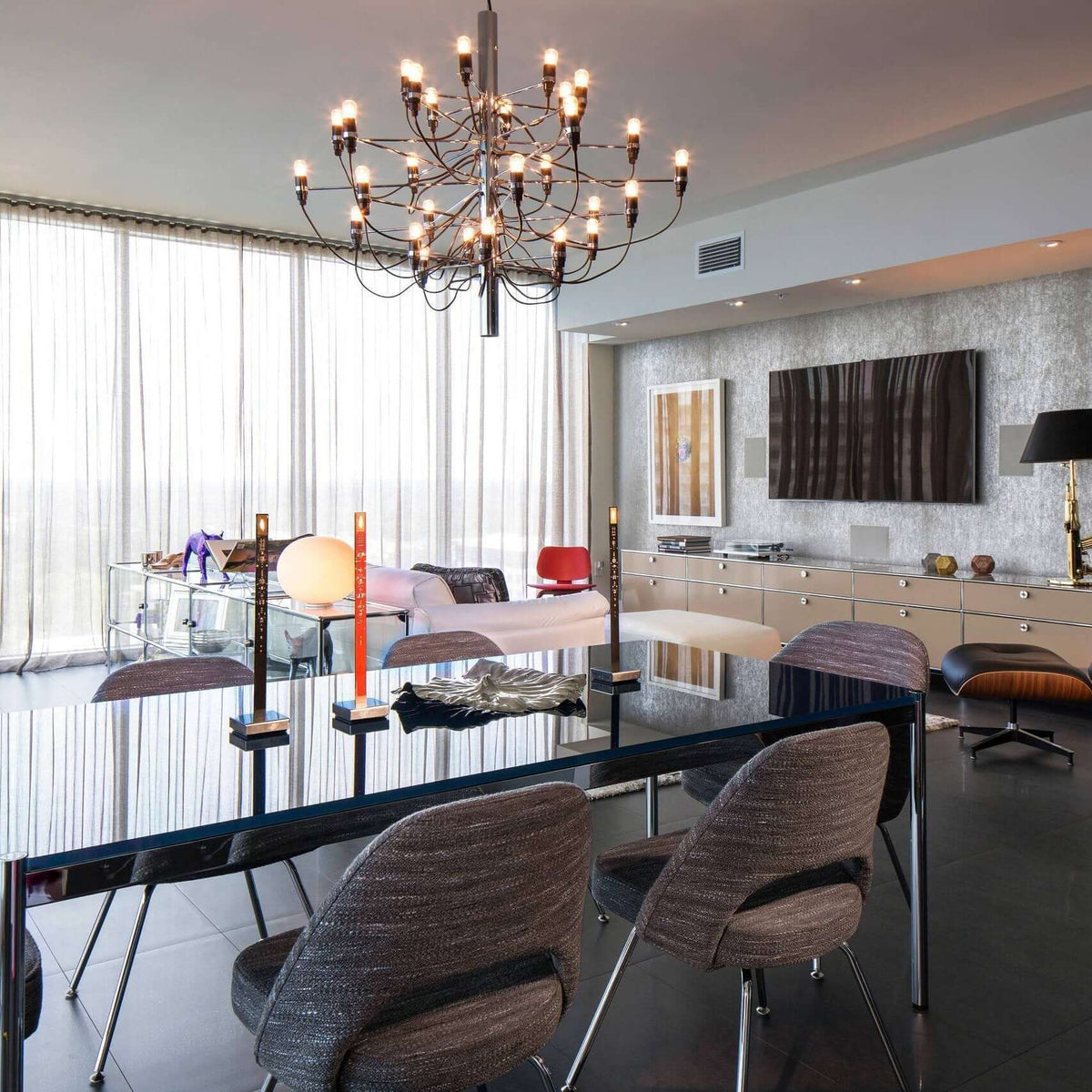 This Clérachic chandelier lights a long glossy dining table in an open-plan room, with sheer curtains and a sideboard behind.