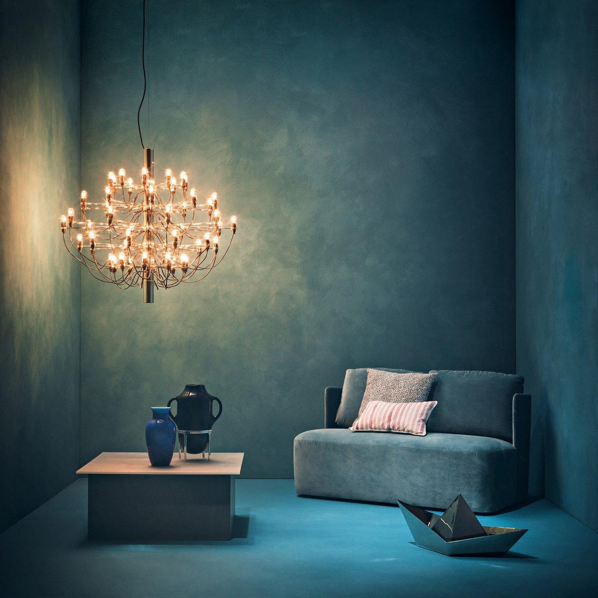 This lit Clérachic chandelier hangs in a teal-painted room above a low table, a grey sofa and two blue vases.