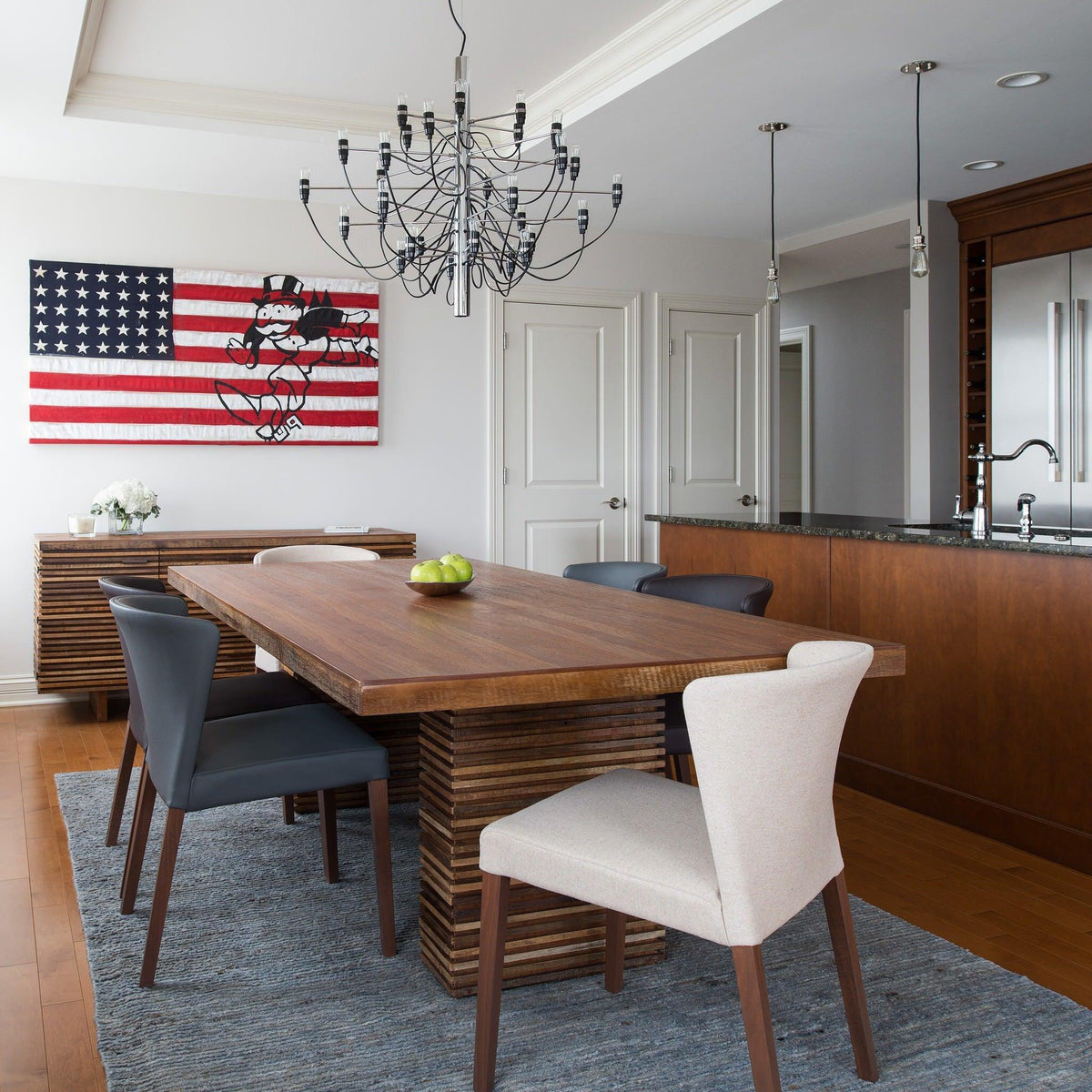 A Clérachic chandelier hangs over a wooden dining table with grey and cream chairs, a stars-and-stripes artwork on the wall behind.
