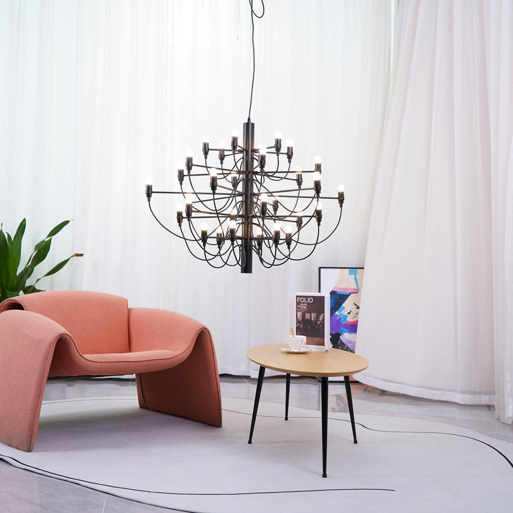 This matt black Clérachic chandelier hangs behind a coral armchair and a round wooden table, in front of white curtains and a plant.