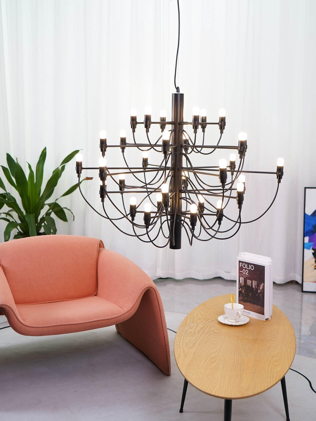 A matt black Clérachic chandelier hangs above a coral armchair and an oval table with a book and cup, beside a leafy plant.