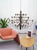 A matt black Clérachic chandelier hangs above a coral armchair and an oval table with a book and cup, beside a leafy plant.