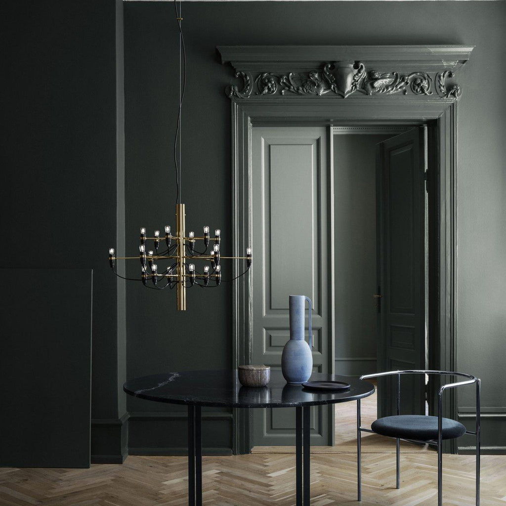 This Clérachic chandelier has a brass stem and hangs in a dark green-grey room beside tall panelled doors and a black round table.