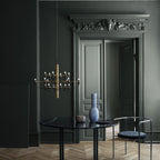 This Clérachic chandelier has a brass stem and hangs in a dark green-grey room beside tall panelled doors and a black round table.