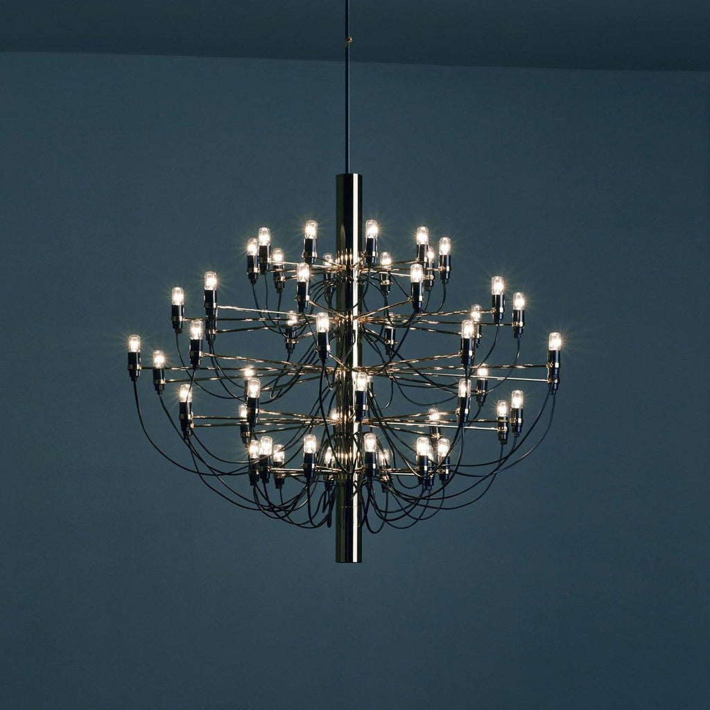 A lit Clérachic tiered chandelier hangs against a deep blue wall, its slim arms and bulbs glowing.
