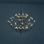 A lit Clérachic tiered chandelier hangs against a deep blue wall, its slim arms and bulbs glowing.