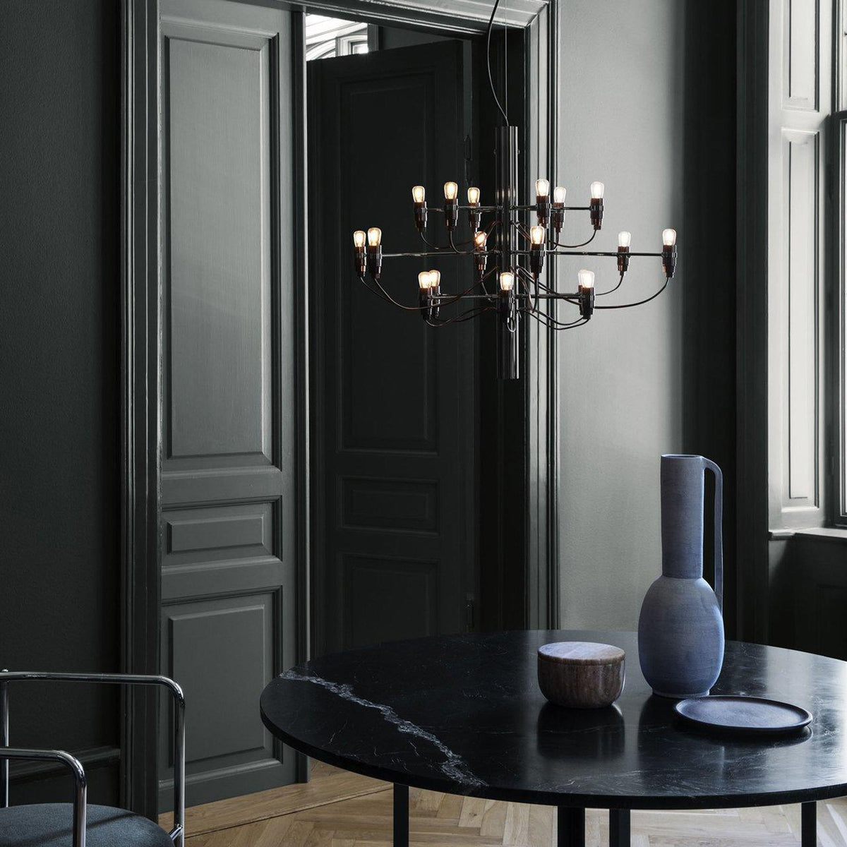 A Clérachic chandelier hangs near dark panelled doors, above a black marble table with a tall blue vase and a bowl.